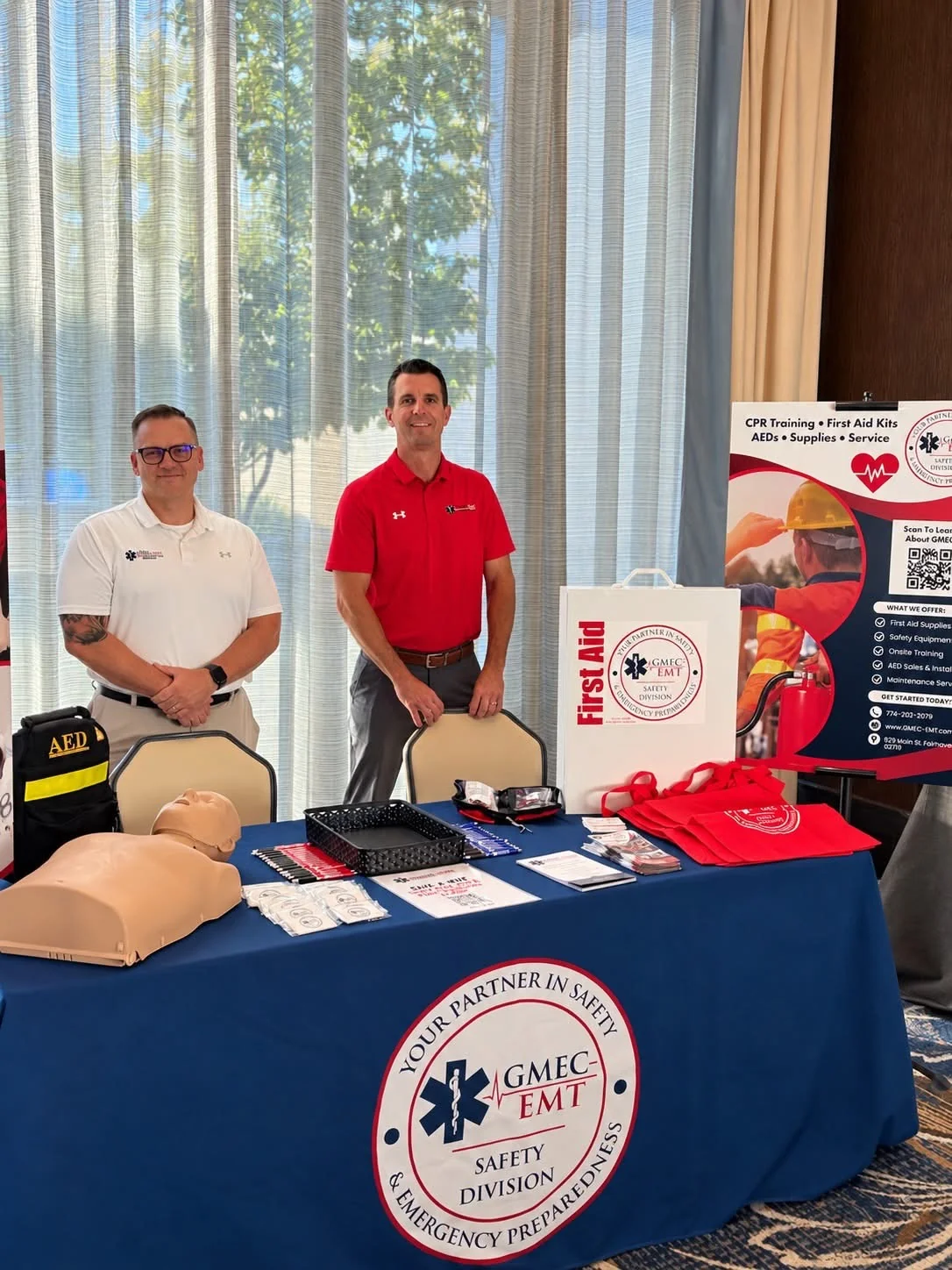 Two GMEC representatives at a safety training display with a CPR mannequin, AED and first aid supplies.