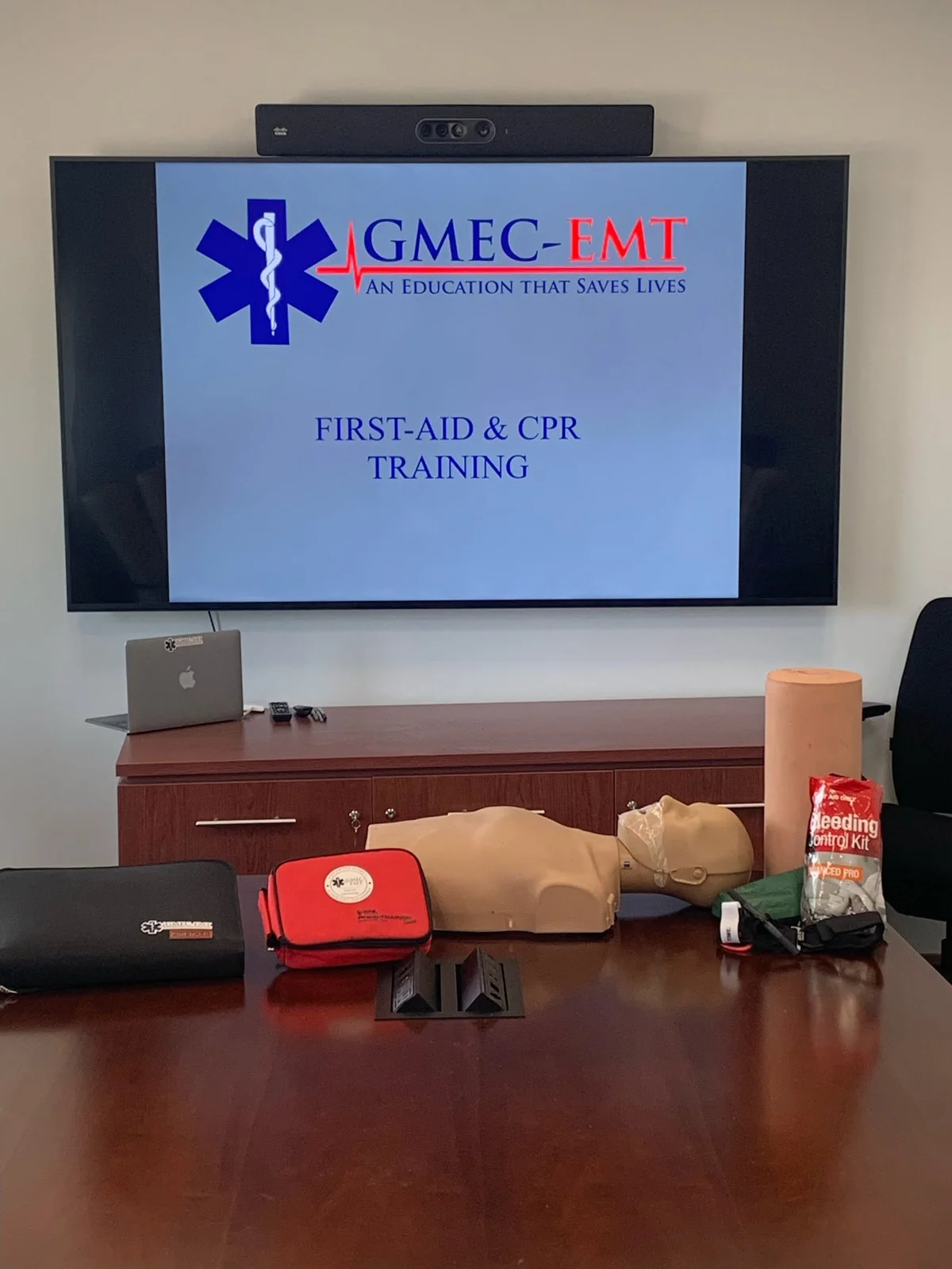 GMEC first-aid and CPR training setup with a manikin, AED trainer and bleeding-control equipment.